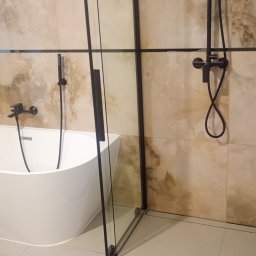 Kris-Rem Remont i Wykończenia wnętrz - Nowoczesna łazienka z owalną białą wanną i czarną, szklaną kabiną prysznicową typu walk-in z czarną armaturą na beżowych płytkach imitujących marmur.