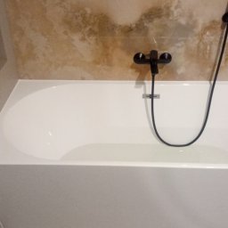 Kris-Rem Remont i Wykończenia wnętrz - Nowoczesna, prostokątna wanna wypełniona wodą, z czarną armaturą i prysznicem na tle beżowych płytek imitujących marmur, widoczna półka w ścianie z geometrycznym wzorem.