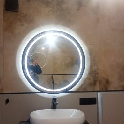 Kris-Rem Remont i Wykończenia wnętrz - Okrągłe lustro z podświetleniem LED zawieszone nad białą umywalką z czarną baterią, na ścianie płytki imitujące kamień w odcieniach beżu i brązu, widoczne odbicie osoby robiącej zdjęcie.