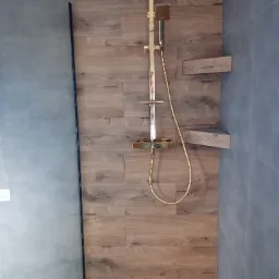 Nowoczesna kabina prysznicowa z deszczownicą w kolorze złotym, obudowana płytkami imitującymi drewno, z p&oacute;łkami narożnymi i odpływem liniowym.