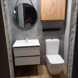 Nowoczesna łazienka z umywalką na szafce z dwiema szufladami, wiszącą szafką z drewnianym frontem, okrągłym lustrem na pasku i toaletą, szare płytki na ścianach i drewnopodobna podłoga, widoczne...