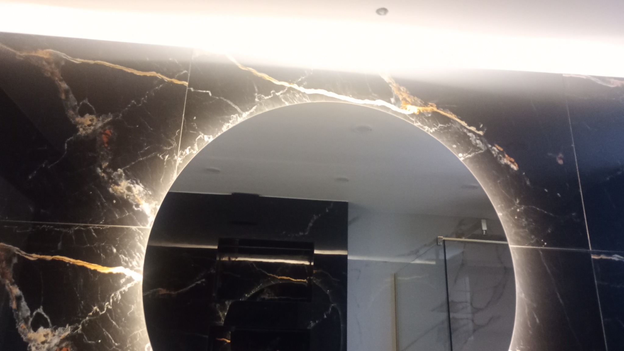 Czarne płytki imitujące marmur z wyraźnym złotym użyleniem, okrągłe podświetlane lustro odbijające fragment kabiny prysznicowej, sufit z oświetleniem LED.