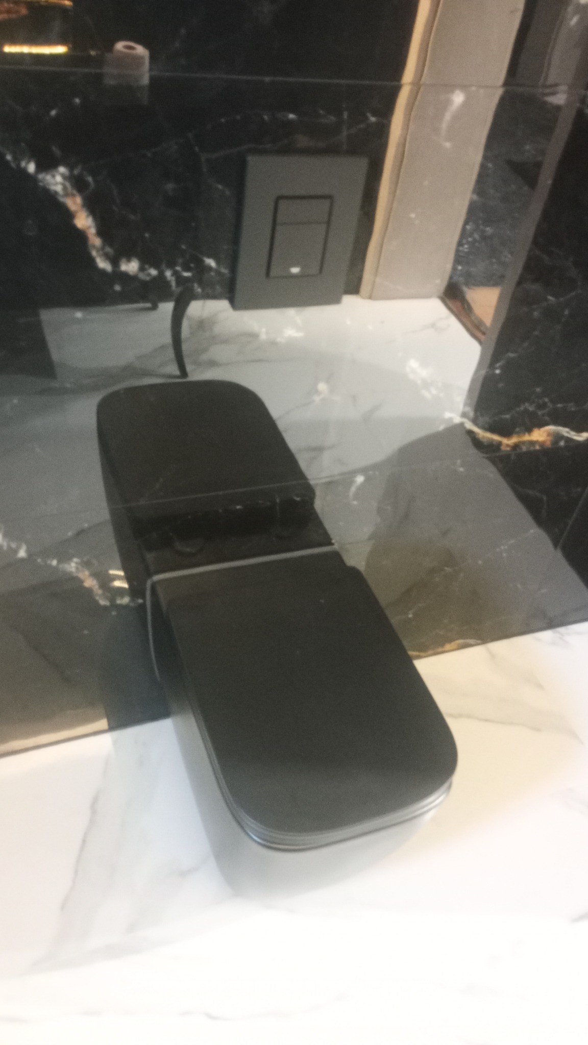 Czarny, nowoczesny przycisk spłuczki toalety zamontowany na ścianie z czarnego marmuru z białymi żyłami, nad białą marmurową umywalką.