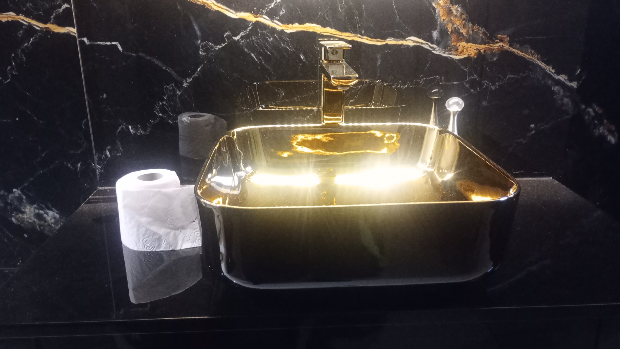 Czarna umywalka nablatowa w kształcie prostokąta z pozłacaną armaturą i czarnym marmurem w tle, obok rolka papieru toaletowego.