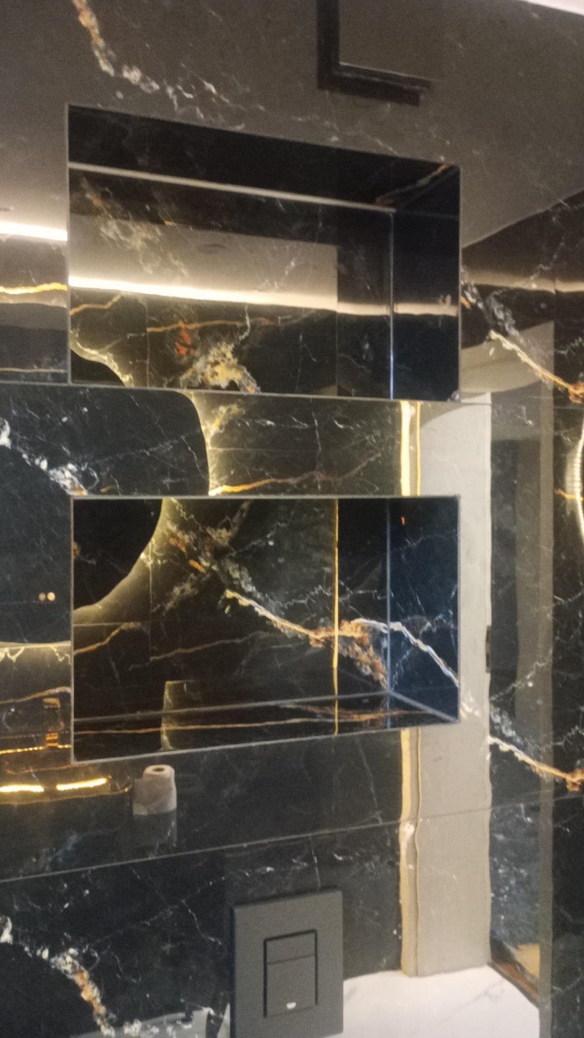 Czarne marmurowe płytki z złotymi żyłami tworzą półki we wnęce, podświetlone od góry, z czarnym włącznikiem światła na ścianie i fragmentem armatury.