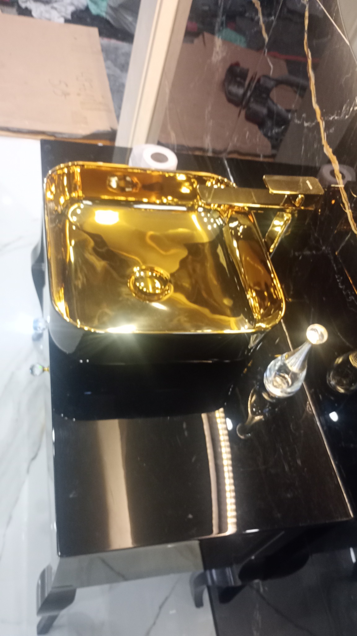 Błyszcząca, złota umywalka nablatowa z dopasowaną armaturą na czarnym blacie z marmurowym wzorem, uzupełnona flakonem perfum.