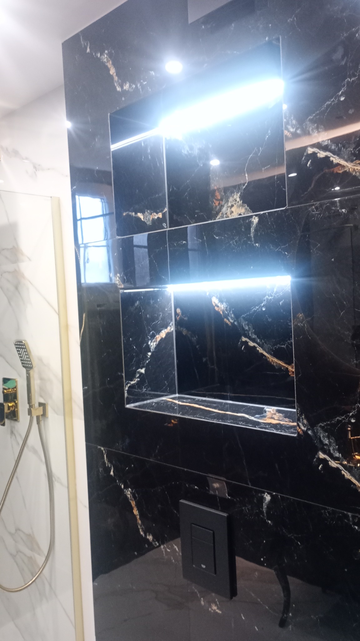 Czarna, marmurkowa ściana kabiny prysznicowej z wnękami oświetlonymi listwą LED, widoczna bateria prysznicowa i czarny włącznik światła.