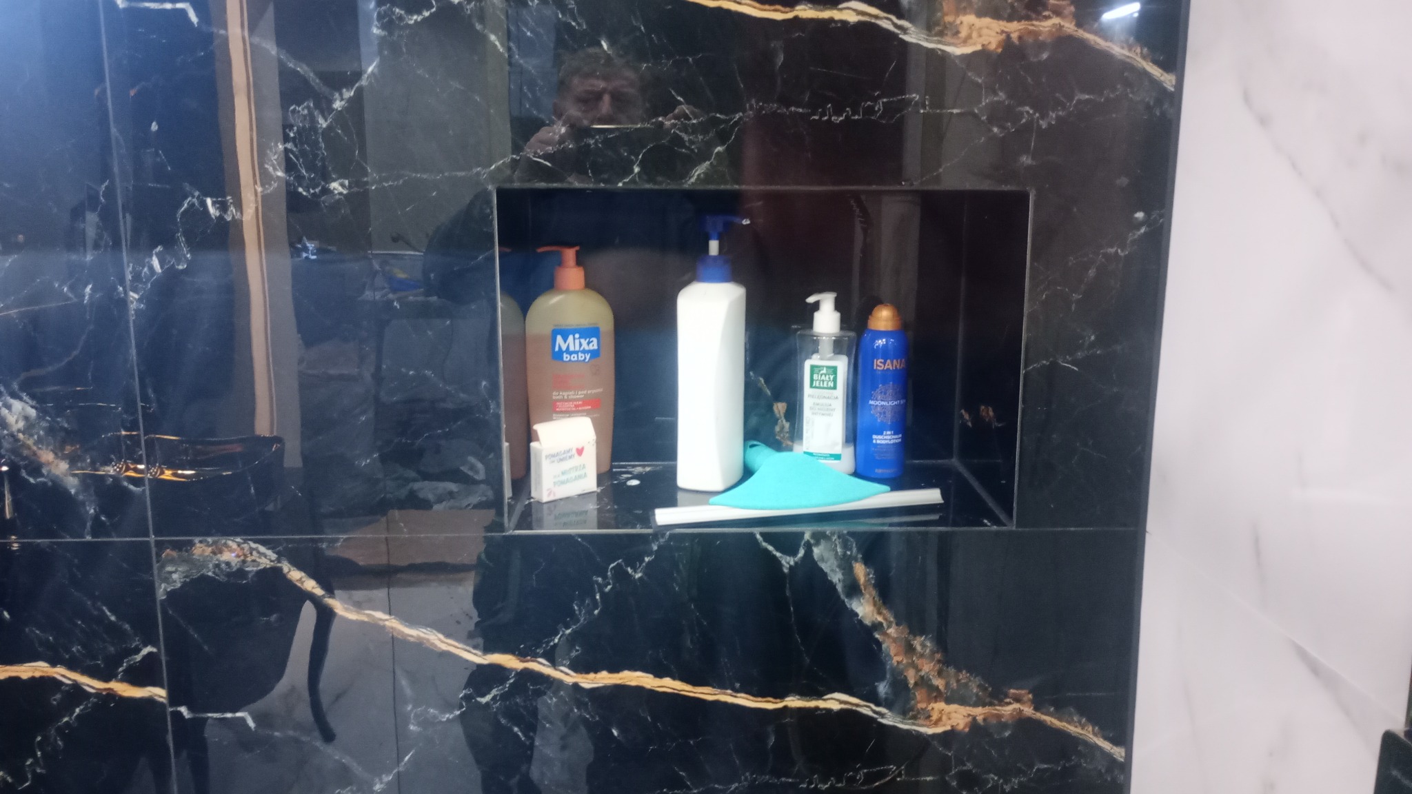 Półka wnękowa w łazience wyłożona czarnymi płytkami imitującymi marmur, z widocznymi kosmetykami i akcesoriami higienicznymi, odbicie twarzy w płytkach.