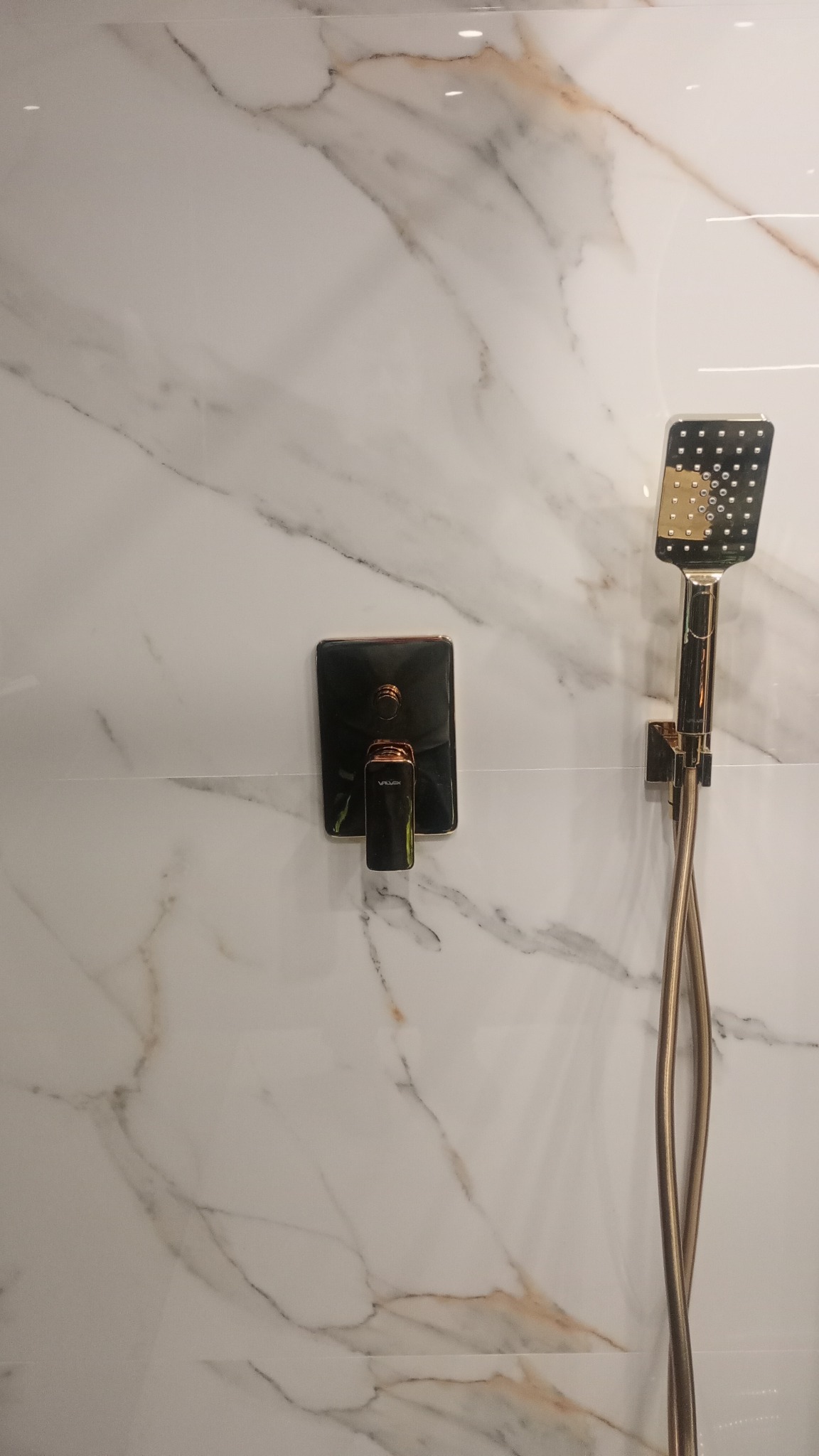 Bateria prysznicowa w kolorze szczotkowanego złota, zamontowana na ścianie wyłożonej białymi płytkami imitującymi marmur z wyraźnym żyłowaniem.