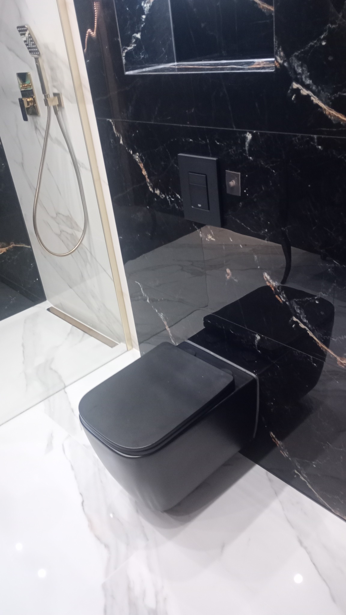 Nowoczesna, czarno-biała łazienka z wiszącą toaletą o matowym wykończeniu, kabiną prysznicową ze złotymi akcentami i marmurowymi płytkami na ścianach i podłodze.