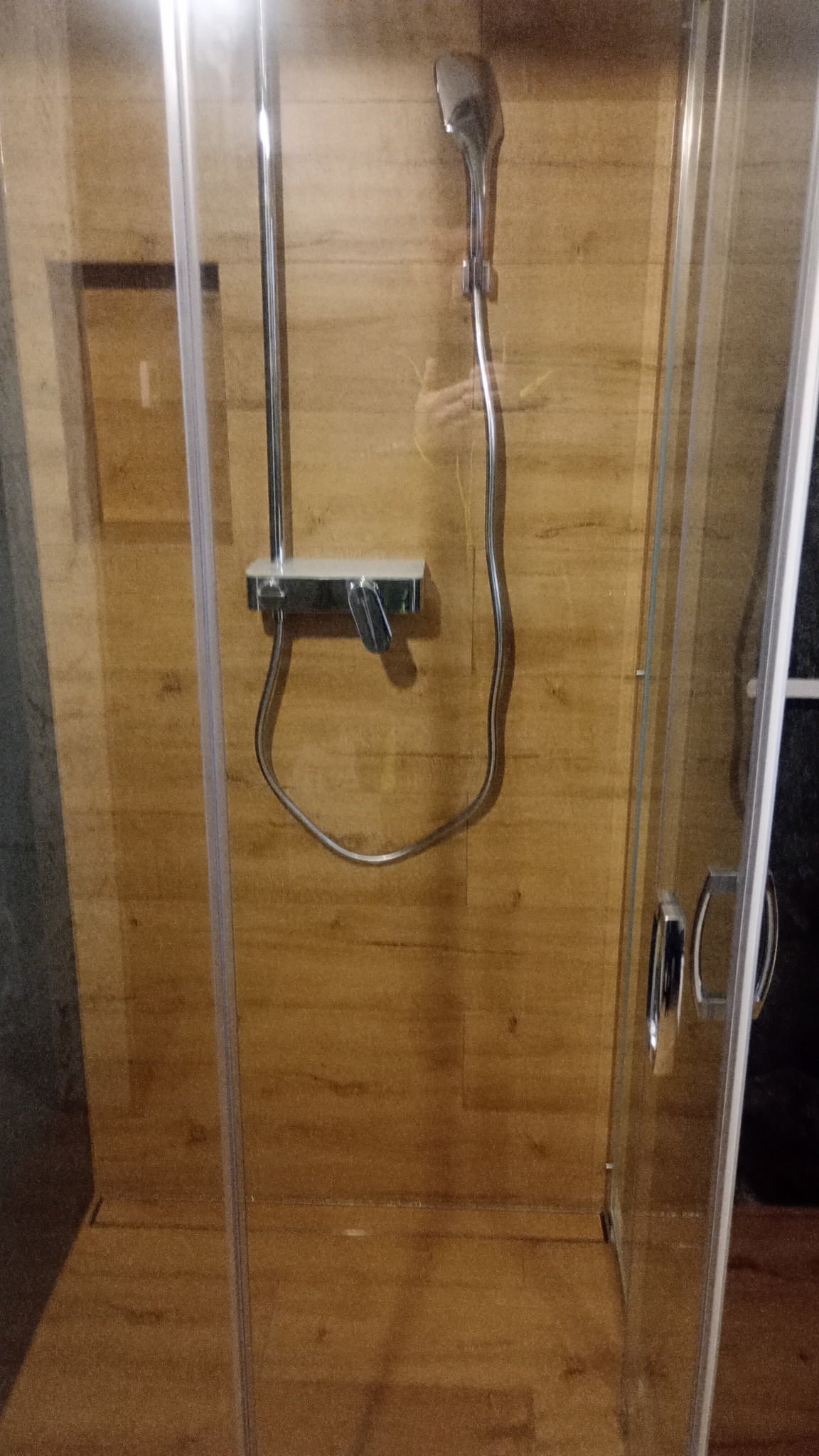 Szklana kabina prysznicowa z chromowaną armaturą na tle drewnopodobnych płytek ściennych i podłogowych.