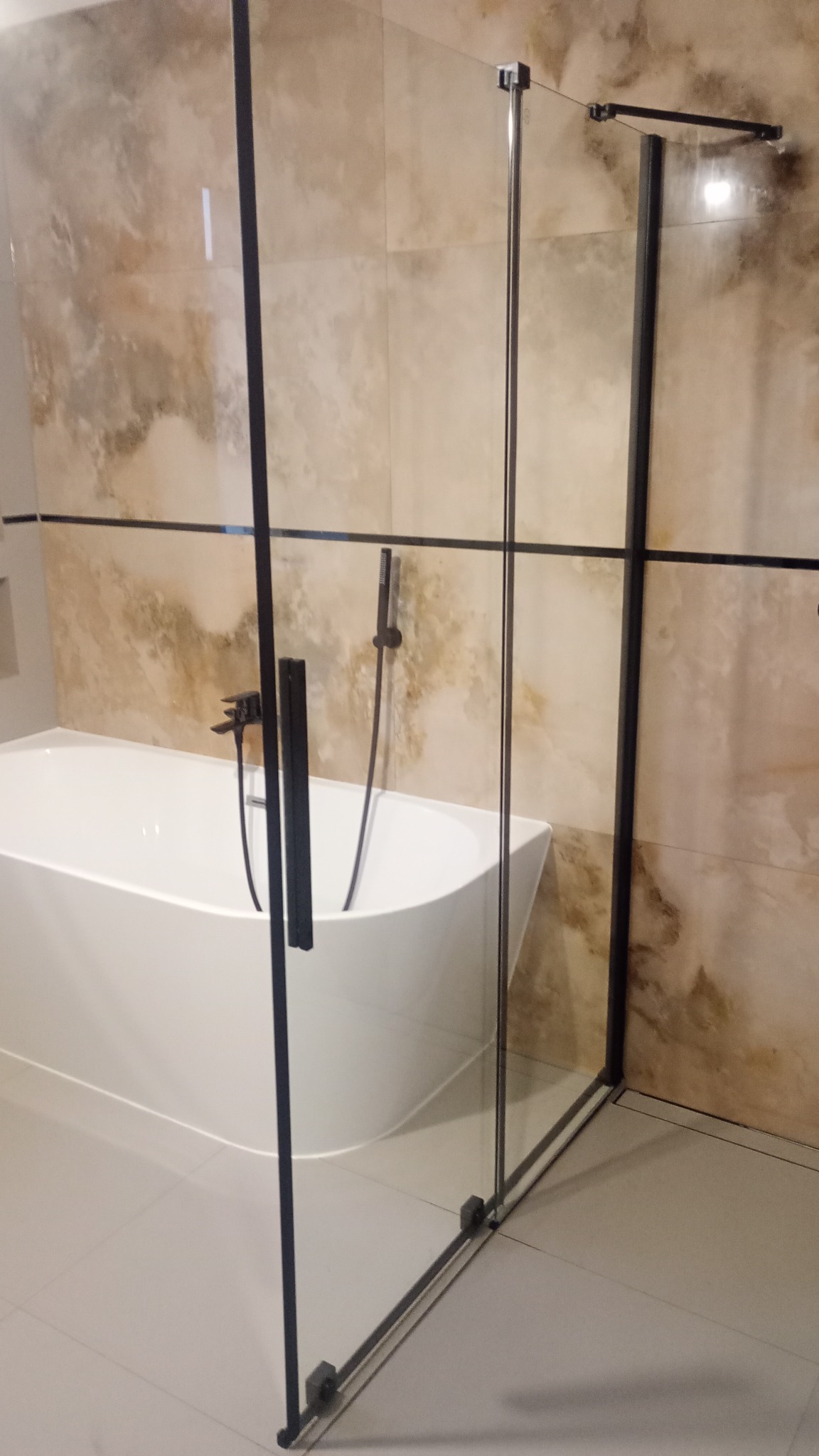 Nowoczesna łazienka z wolnostojącą białą wanną i kabiną prysznicową ze szkła z czarnymi profilami, na tle beżowych płytek imitujących marmur.