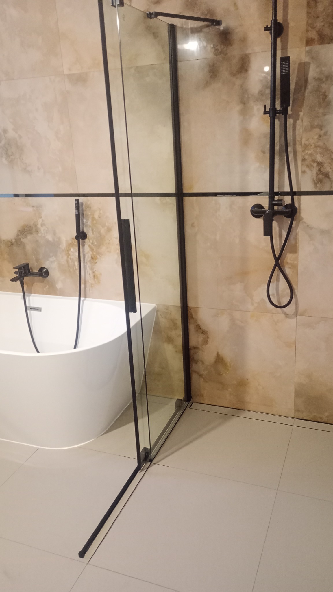 Nowoczesna łazienka z owalną białą wanną i czarną, szklaną kabiną prysznicową typu walk-in z czarną armaturą na beżowych płytkach imitujących marmur.