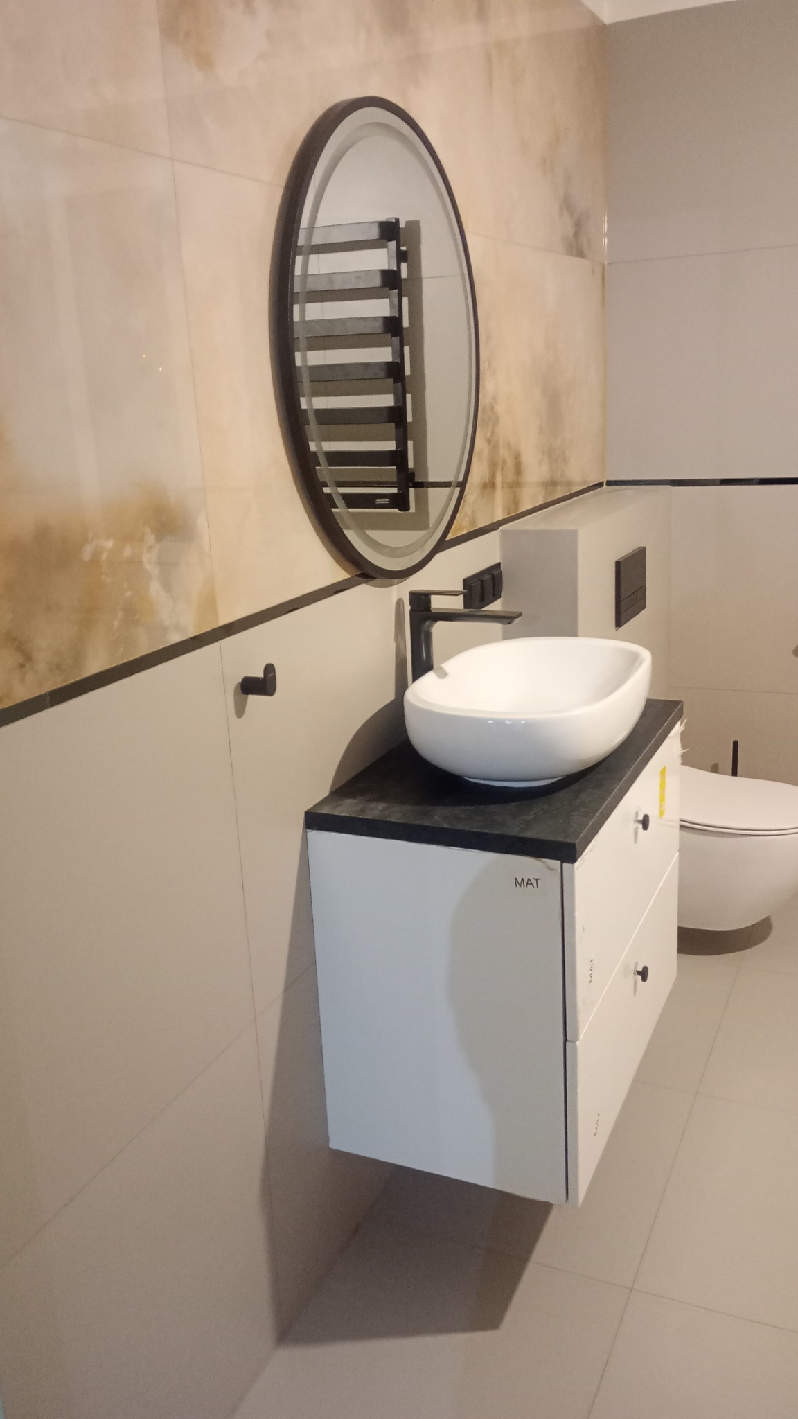 Nowoczesna łazienka z owalną umywalką nablatową, czarną armaturą i okrągłym lustrem z widocznym grzejnikiem drabinkowym w odbiciu, na beżowych płytkach imitujących kamień.