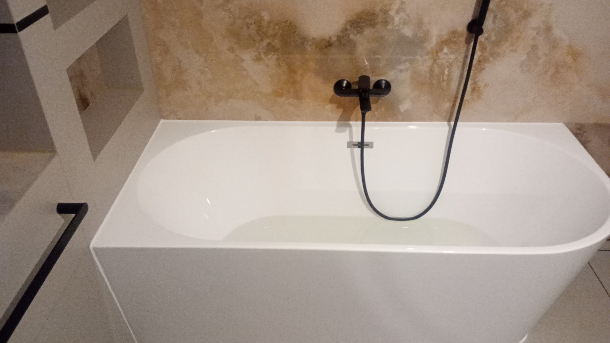 Nowoczesna, prostokątna wanna wypełniona wodą, z czarną armaturą i prysznicem na tle beżowych płytek imitujących marmur, widoczna półka w ścianie z geometrycznym wzorem.