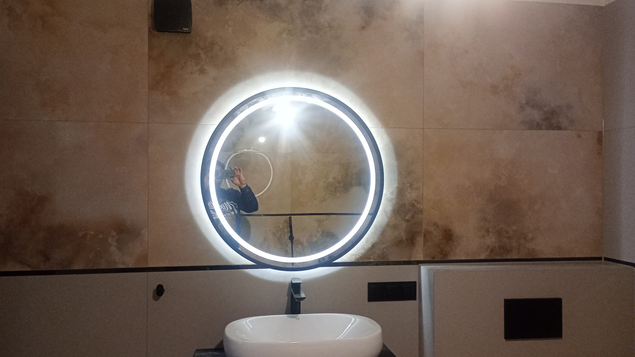 Okrągłe lustro z podświetleniem LED zawieszone nad białą umywalką z czarną baterią, na ścianie płytki imitujące kamień w odcieniach beżu i brązu, widoczne odbicie osoby robiącej zdjęcie.