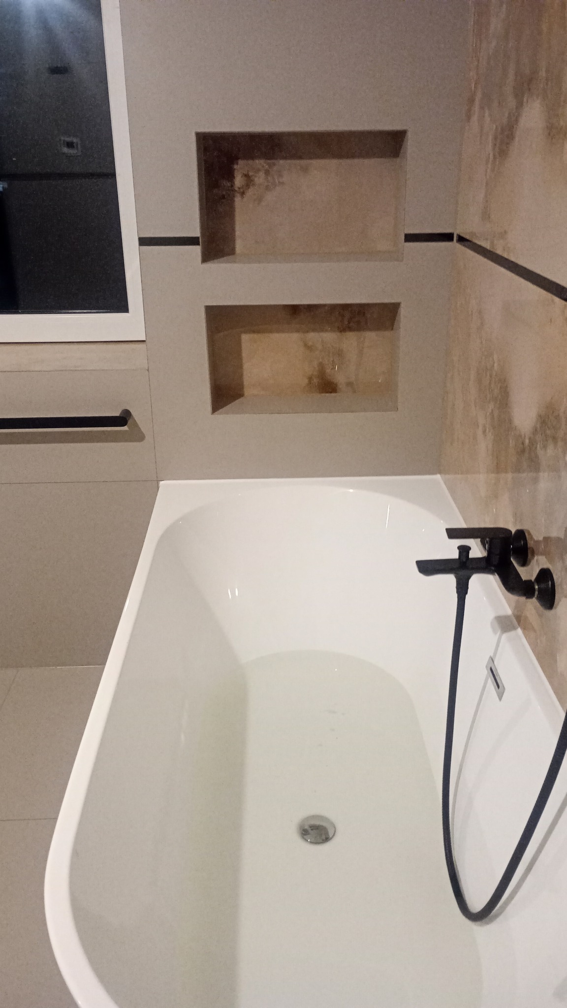 Biała wanna z czarną armaturą, w tle beżowe płytki z wnękami, minimalistyczny design łazienki.