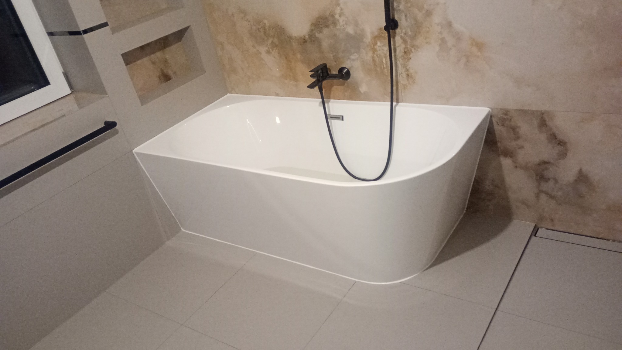 Nowoczesna biała wanna wolnostojąca z czarną armaturą na tle beżowych płytek imitujących marmur, widoczny odpływ liniowy w podłodze.