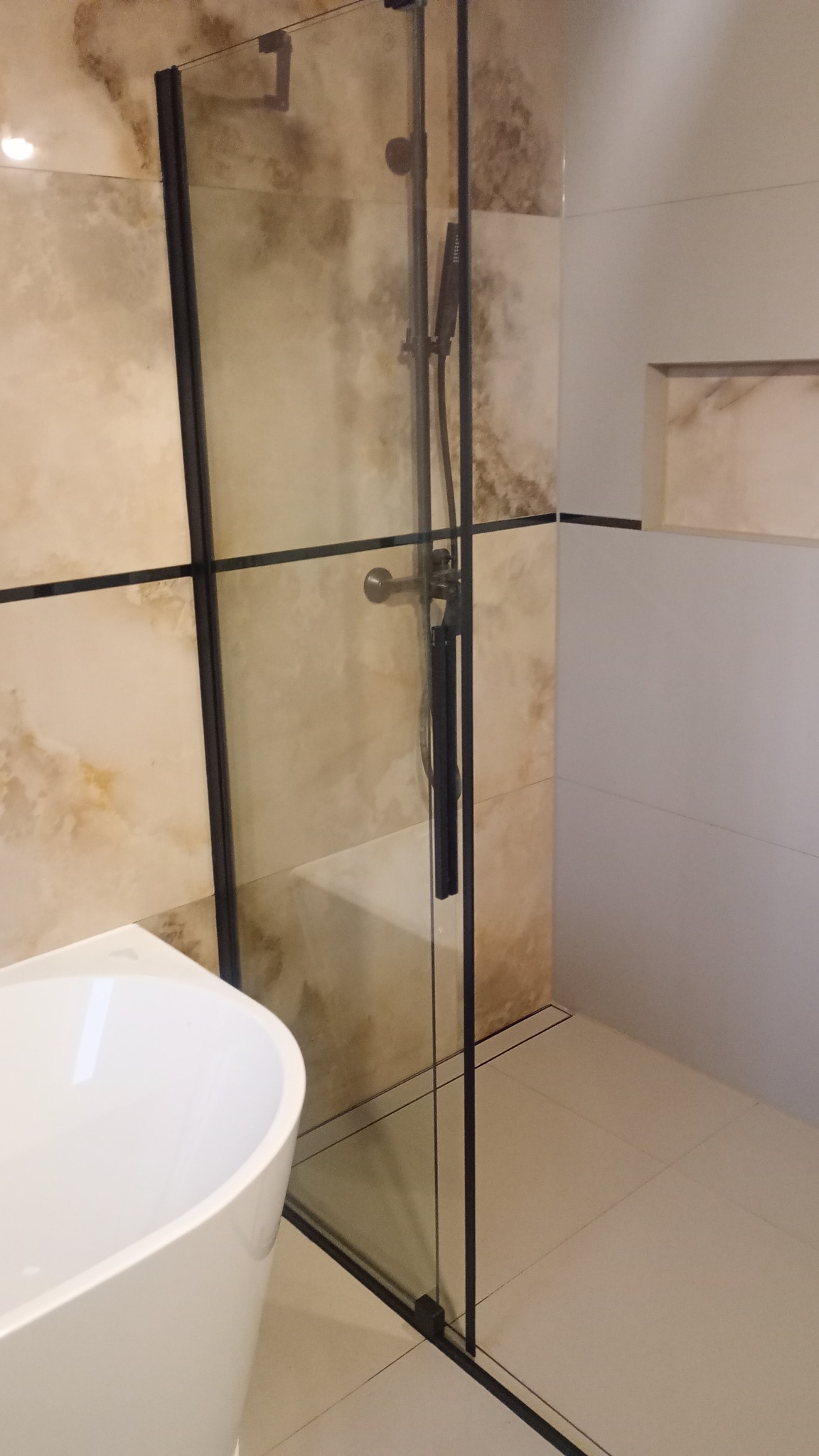 Nowoczesna kabina prysznicowa z czarnymi profilami i deszczownicą, obok wolnostojąca wanna i beżowe płytki imitujące marmur