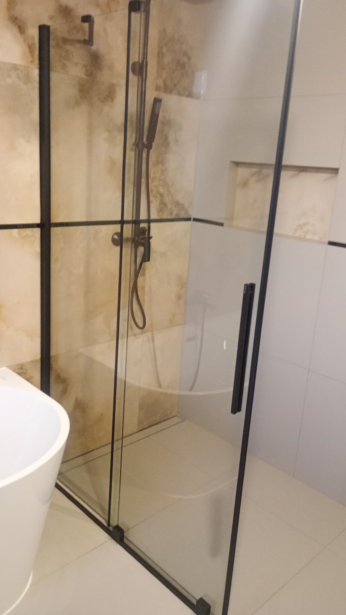 Nowoczesna kabina prysznicowa z czarnymi profilami i deszczownicą, widoczna umywalka nablatowa, beżowe i białe płytki na ścianach.