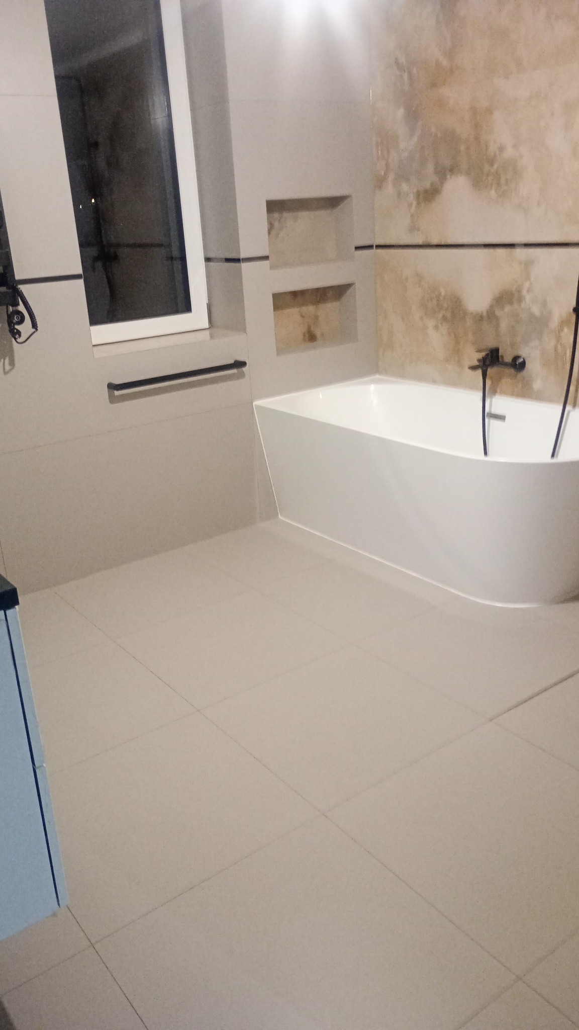 Nowoczesna łazienka z białą wanną wolnostojącą, beżowymi płytkami na podłodze i ścianach, wnękami w ścianie oraz czarną armaturą.