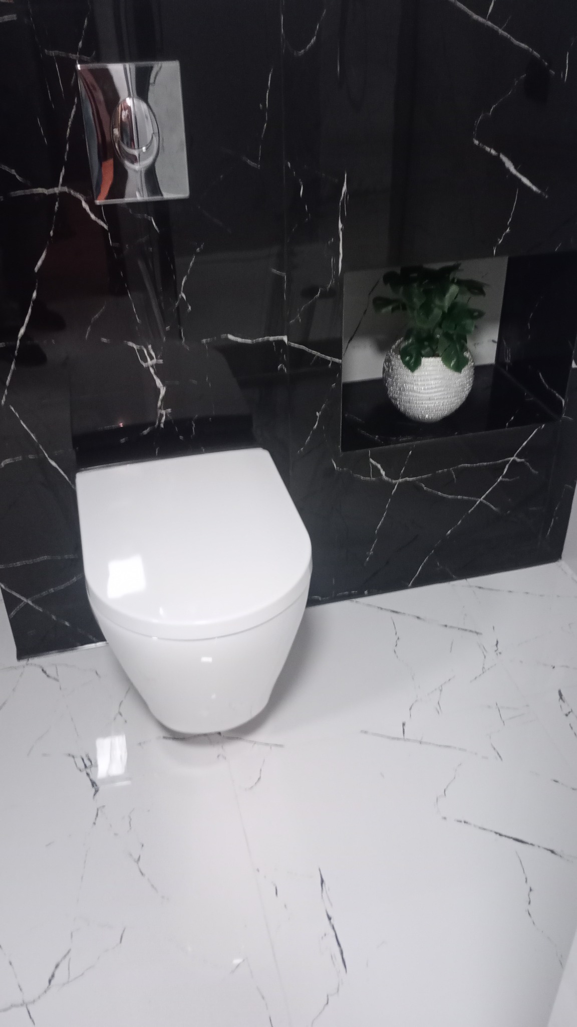 Nowoczesna toaleta podwieszana w łazience wykończonej czarno-białym marmurem z żyłami; na ścianie wnęka z dekoracyjną rośliną w srebrnej doniczce.