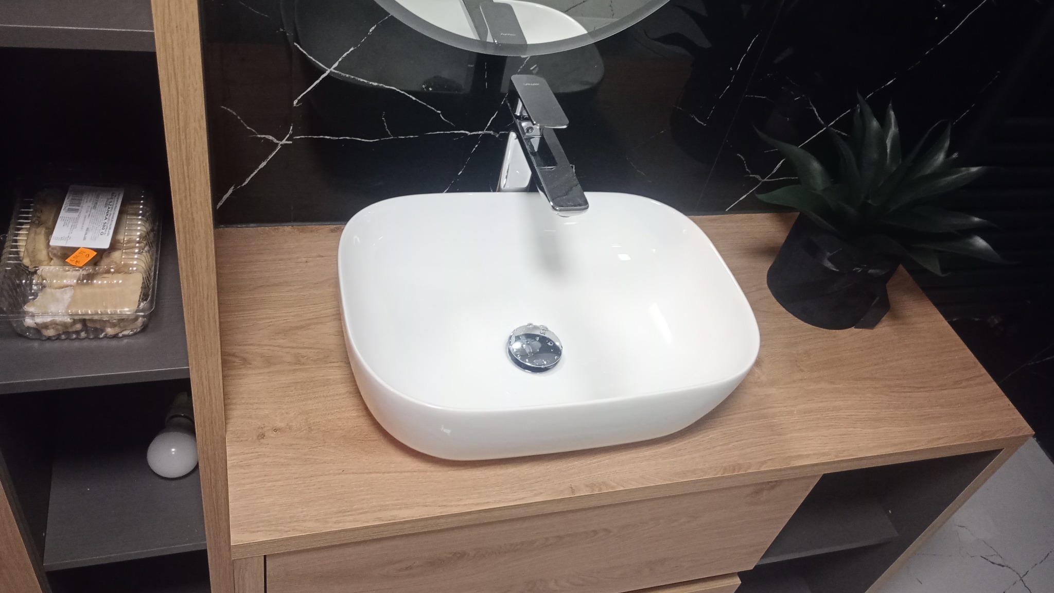 Biała, owalna umywalka nablatowa z chromowaną armaturą na drewnopodobnym blacie szafki łazienkowej, z czarnym marmurem na ścianie i sztuczną rośliną w tle, widok z góry.