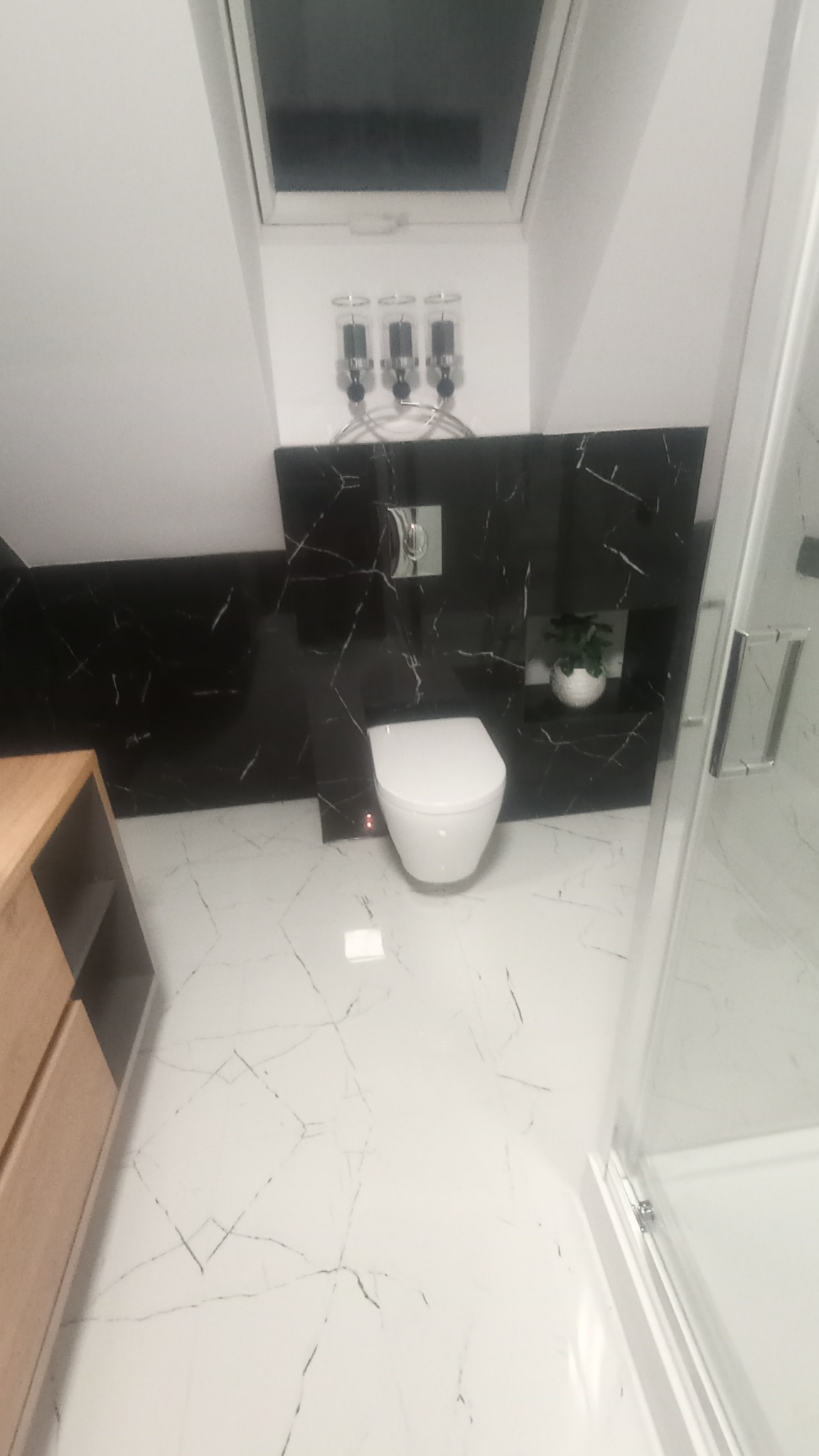 Nowoczesna łazienka na poddaszu z białą armaturą, czarnymi płytkami imitującymi marmur na ścianie i białą podłogą z czarnymi żyłkami, oświetlona światłem z okna dachowego.