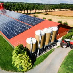 Widok z lotu ptaka na budynek gospodarczy z czerwonym dachem pokrytym panelami słonecznymi, obok silosy i czerwony traktor na tle zieleni i pól uprawnych.