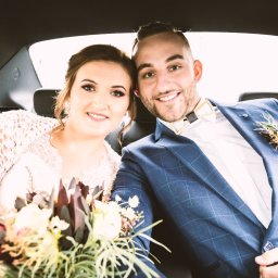 DS. PRO STUDIO - Uśmiechnięta para młoda siedzi w samochodzie, panna młoda trzyma bukiet z ciemnymi proteami, pan młody w niebieskiej kraciastej marynarce z butonierką.