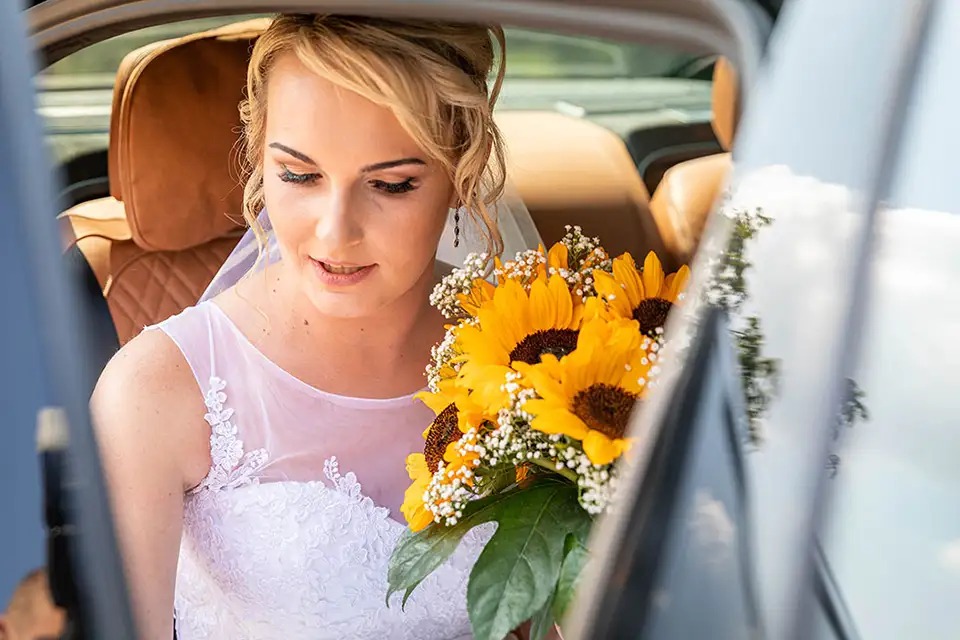 Panna młoda w sukni ślubnej z bukietem słoneczników siedząca w samochodzie z brązową skórzaną tapicerką.