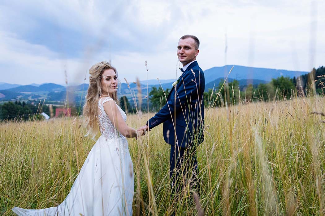 Para młoda trzymająca się za ręce na tle łąki i gór w pochmurny dzień; panna młoda w białej sukni z koronkowym wykończeniem, pan młody w granatowym garniturze.