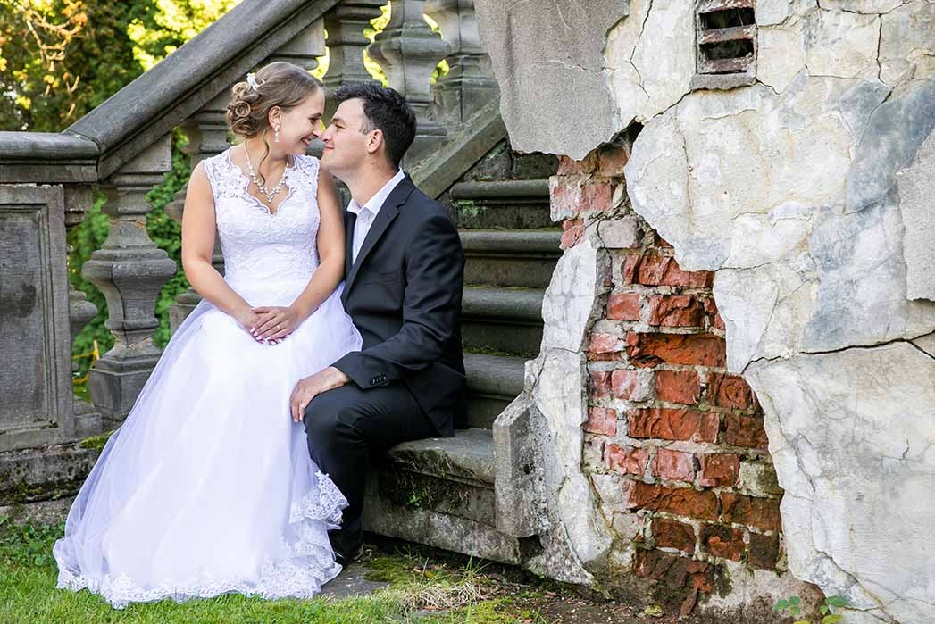 Para młoda, panna młoda w koronkowej sukni i pan młody w czarnym garniturze, siedzą na starych kamiennych schodach obok ściany z cegły z odpadającym tynkiem.