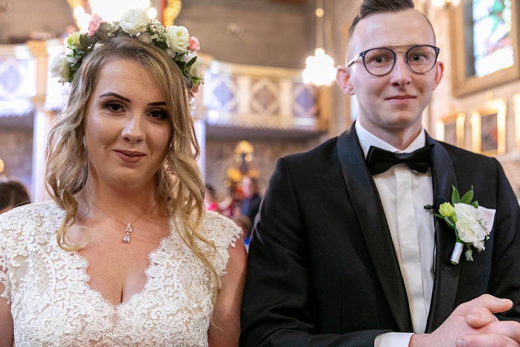 Para młoda w kościele: Panna młoda w koronie z kwiatów i sukni z koronki, Pan młody w czarnym smokingu z muchą i butonierką.