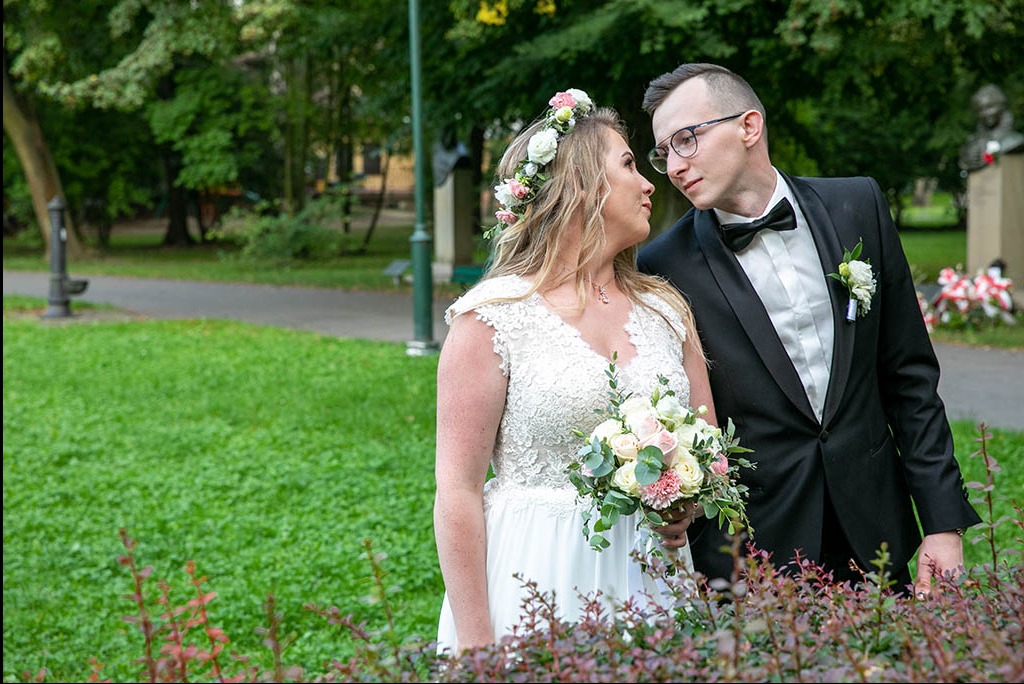 Para młoda w parku: Panna młoda w koronkowej sukni i wianku, trzyma bukiet, pan młody w czarnym smokingu z muchą, oboje patrzą na siebie z uśmiechem.