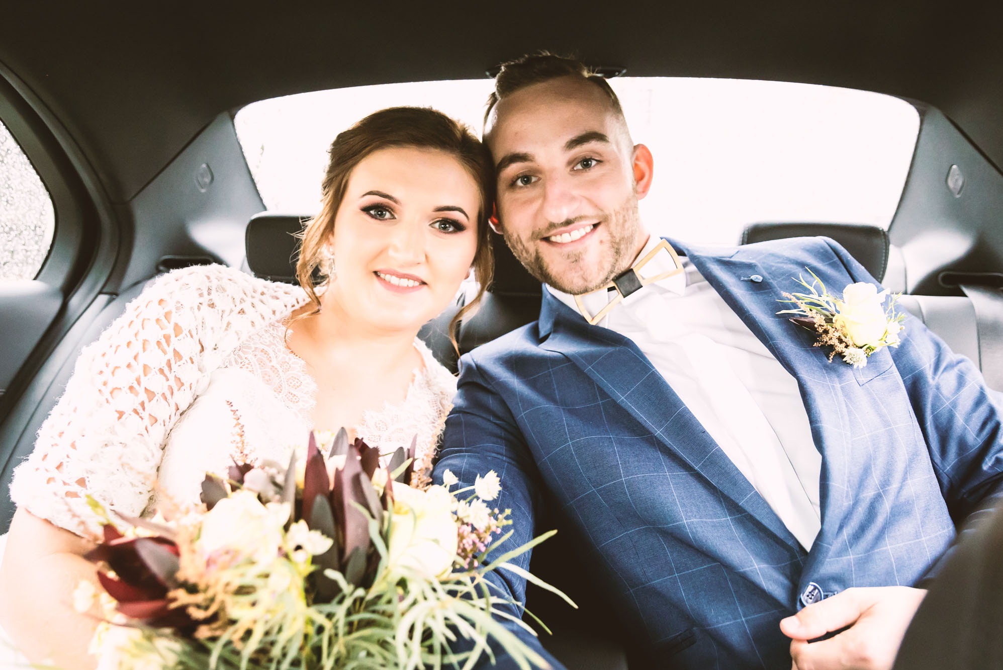 Uśmiechnięta para młoda siedzi w samochodzie, panna młoda trzyma bukiet z ciemnymi proteami, pan młody w niebieskiej kraciastej marynarce z butonierką.