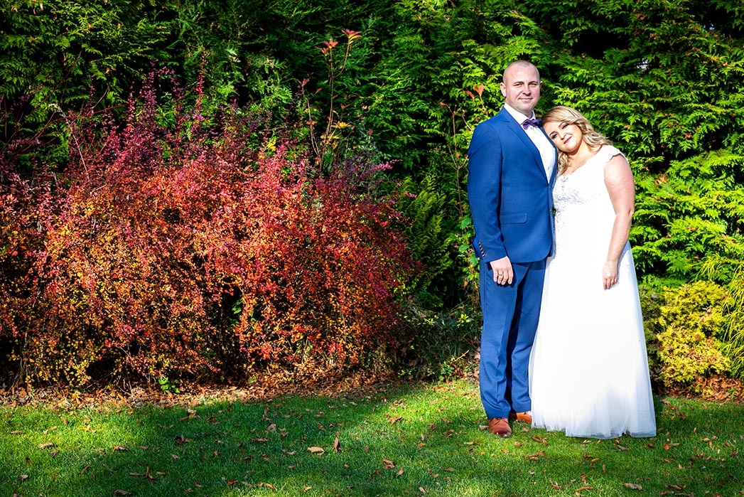 Para młoda: Panna młoda w białej sukni ślubnej opiera głowę na ramieniu pana młodego w niebieskim garniturze z fioletową muchą na tle zieleni.