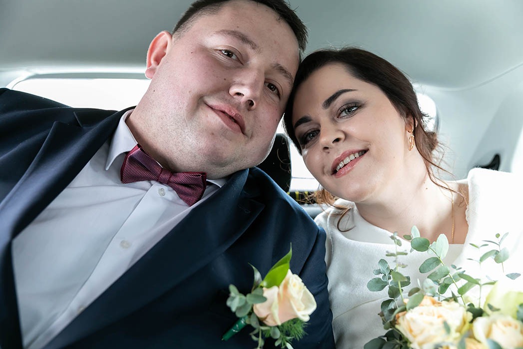 Uśmiechnięta para młoda w samochodzie, zbliżenie na twarze, bukiet kwiatów i bordowa mucha.
