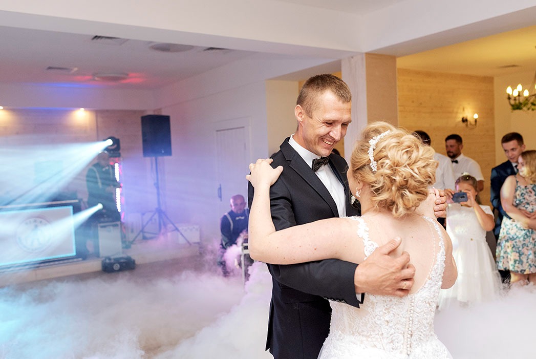 Para młoda w pierwszym tańcu, otoczona sztuczną mgłą, w tle DJ i goście, oświetlenie sceniczne.