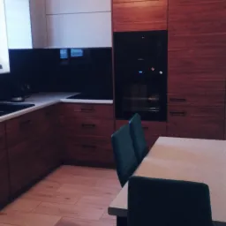 Nowoczesna kuchnia z drewnianymi frontami, czarnym panelem nad blatem i drewnianą podłogą, uzupełniona zielonymi krzesłami przy stole.