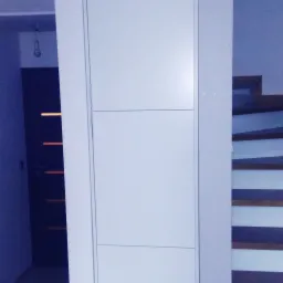 Białe, minimalistyczne drzwi wewnętrzne z poziomymi frezowaniami, zamontowane w jasnym pomieszczeniu z widocznymi schodami i fragmentem ciemnych drzwi w tle.