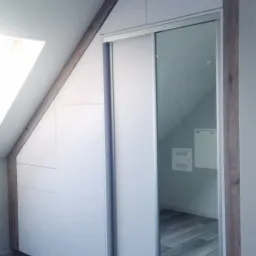 Biała szafa wnękowa z przesuwnymi drzwiami z lustrem, dopasowana do skosu poddasza, z drewnianym wykończeniem.