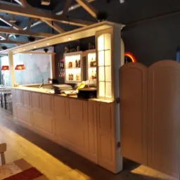 Długa, beżowa lada barowa z szafkami i półkami podświetlonymi od wewnątrz, widoczne wahadłowe drzwi i elementy konstrukcji sufitu w lokalu gastronomicznym.