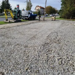 Utwardzanie drogi kruszywem: walec drogowy ugniata szarą nawierzchnię, obok pracownik z łopatą, w tle mini koparka i ciężarówka.