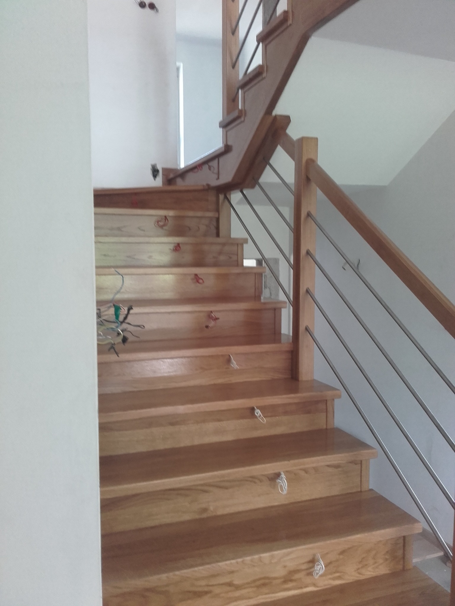 Drewniane schody z metalową balustradą w trakcie montażu, widoczne kable elektryczne i uchwyty montażowe na stopniach.