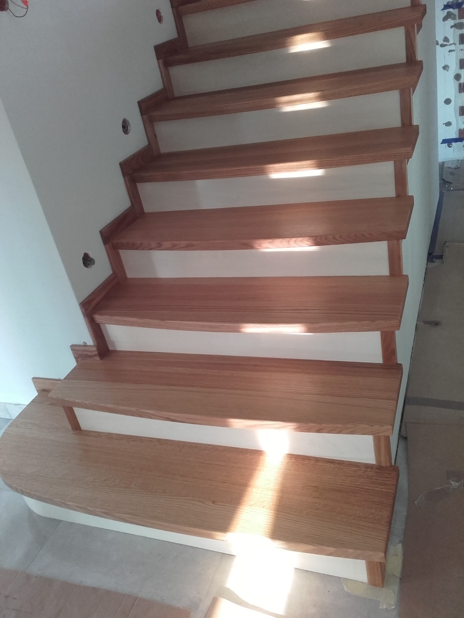 Drewniane schody z jasnego drewna, z zaokrąglonym stopniem początkowym, prowadzące w górę wzdłuż białej ściany z widocznymi otworami na gniazdka elektryczne.