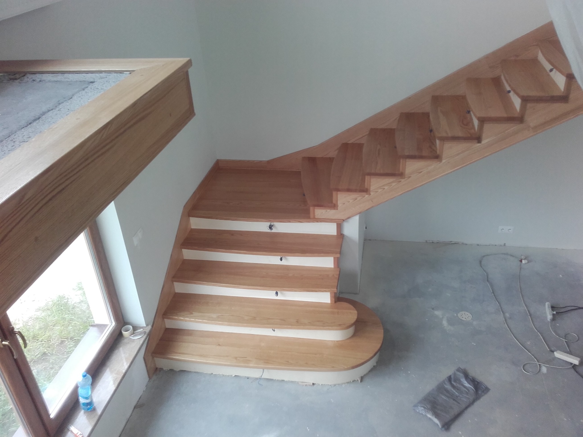 Drewniane schody z zaokrąglonym stopniem początkowym, widok z góry, surowy stan wykończenia podłogi, okno z drewnianą ramą z widokiem na zieleń.