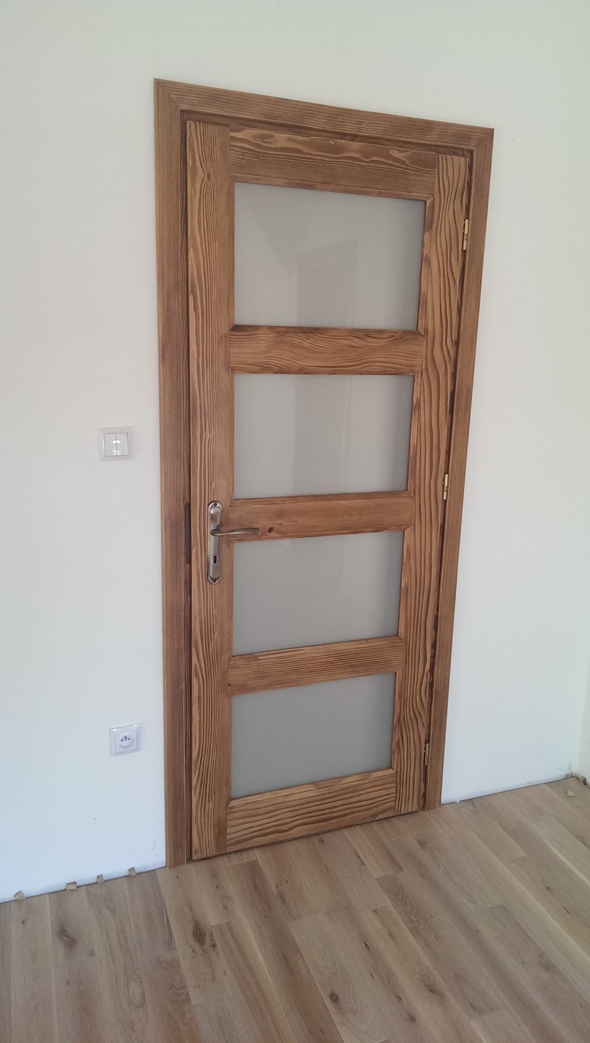 Brązowe, drewniane drzwi wewnętrzne z czterema matowymi szybami, zamontowane w białej ścianie nad parkietem, widoczne gniazdko elektryczne i włącznik światła.