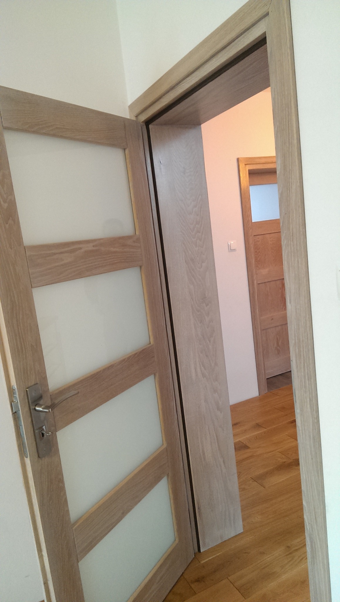 Otwarta drewniana rama drzwiowa z trzema matowymi szybami, widok na parkiet i kolejne drzwi w głębi.