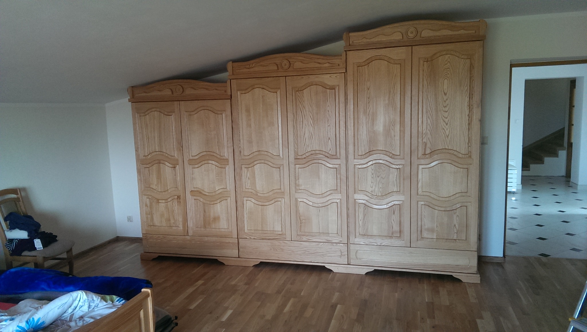 Trzyczęściowa, jasna, drewniana szafa z dekoracyjnymi frezowaniami na frontach, ustawiona w pokoju na poddaszu z drewnianą podłogą, widok na schody w tle.